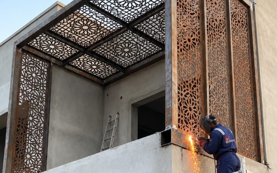 Welder fixing laser-cut steel screens and a pergola roof on a building